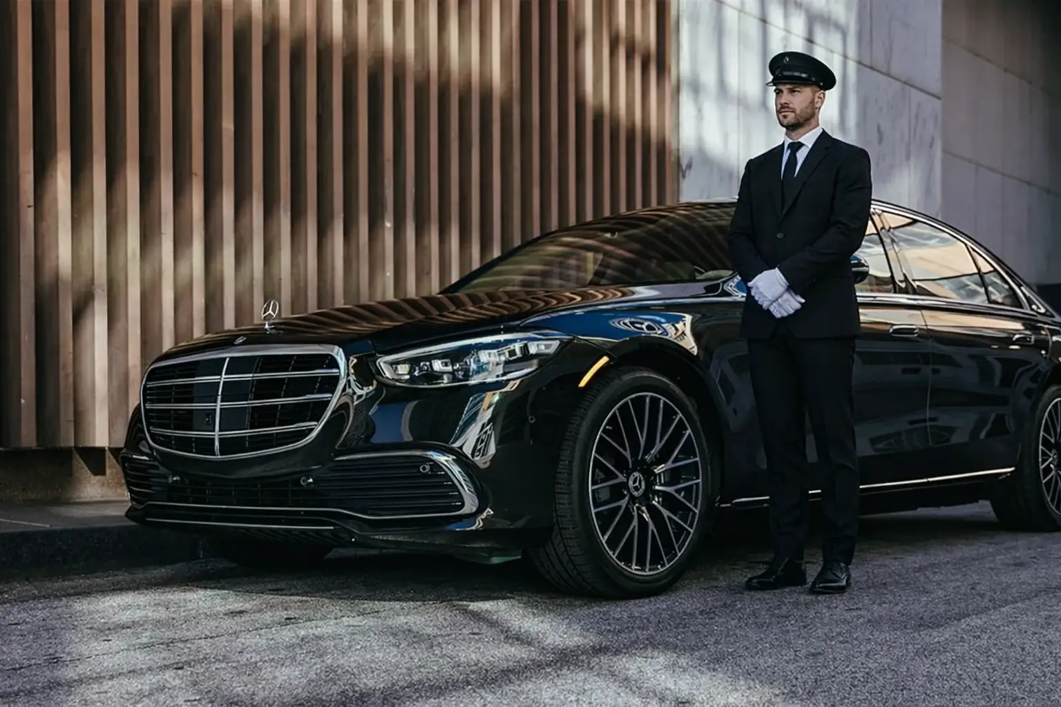 A professional chauffeur in a black suit, cap, and white gloves stands at attention next to a sleek black Mercedes-Benz sedan.
