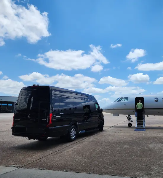 A black Mercedes Sprinter van parked on an airfield next to a private jet under a blue sky with white clouds