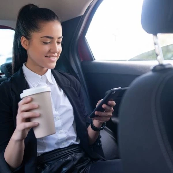 young woman holding coffee using phone inside car